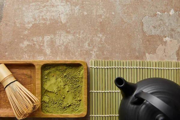 Top view of matcha powder and bamboo whisk on board near black teapot on aged surface — Stock Photo