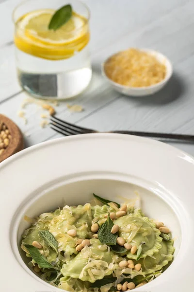 Close up view of green ravioli with pine nuts, grated cheese and sage at wooden table near water with lemon and fork — Stock Photo