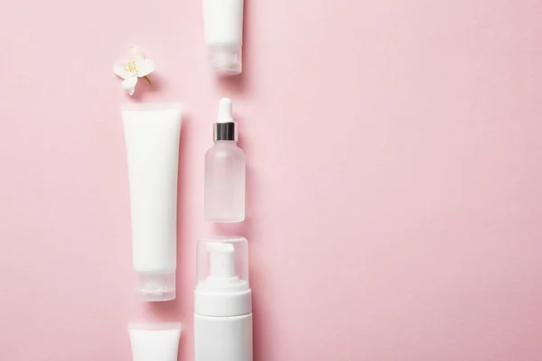 Disposición plana de botella de vidrio cosmético, tubos de crema con crema, dispensador de cosméticos y flor de jazmín en rosa - foto de stock