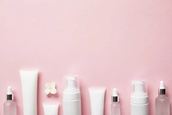 Vista superior de botellas de vidrio cosmético, tubos de crema con crema, dispensadores de cosméticos y flor de jazmín en rosa - foto de stock