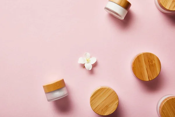Vue du dessus des bocaux éparpillés avec des bouchons crème et en bois, et fleur de jasmin au milieu sur rose — Photo de stock