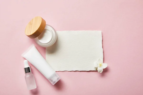 Top view of open jar with cream, cosmetic glass bottle, cream tube, piece of paper and jasmine flower on pink — Stock Photo