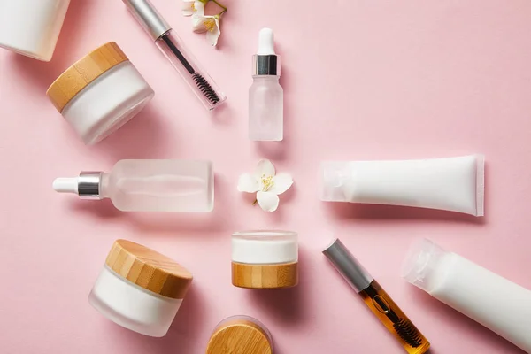Draufsicht auf Gläser, Cremetuben mit Handcreme, Kosmetikglasflaschen, Mascaraflaschen und Jasminblüte in der Mitte auf rosa — Stockfoto