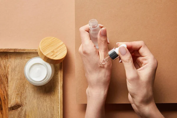 Vista ritagliata di donna in possesso di bottiglia di vetro cosmetico con siero vicino vaso aperto con crema su vassoio di legno su marrone — Foto stock