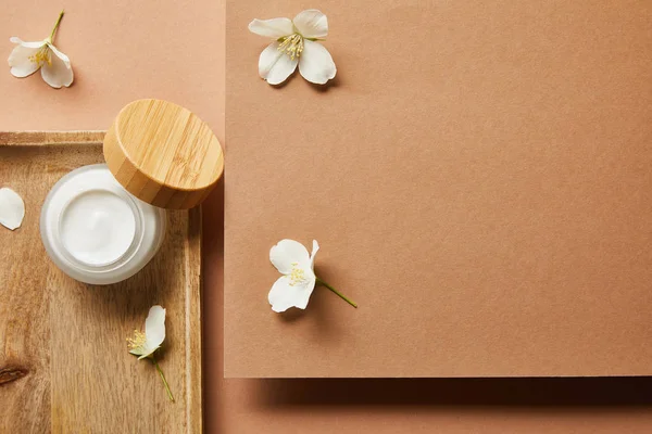 Top view of open jar with cream on wooden tray and scattered jasmine flowers around on brown — Stock Photo