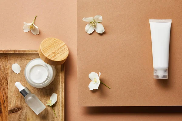 Vista dall'alto di vaso aperto con crema, bottiglia di vetro cosmetico con siero su vassoio di legno, tubo crema e fiori di gelsomino sparsi su marrone — Foto stock