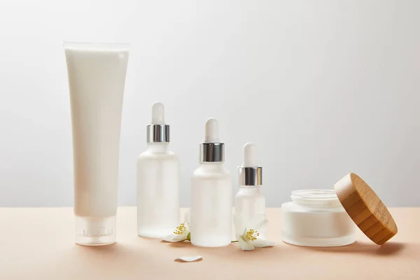 Tube à crème, bouteilles en verre cosmétique, pot ouvert à la crème et quelques fleurs de jasmin sur beige — Photo de stock