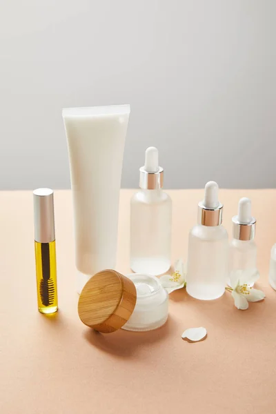 Mascaraflasche mit natürlichem Öl, Cremetubus mit Handcreme, Kosmetikglasflaschen mit Serum, kleines offenes Glas mit Gesichtscreme und Jasminblüte auf beige isoliert auf grau — Stockfoto