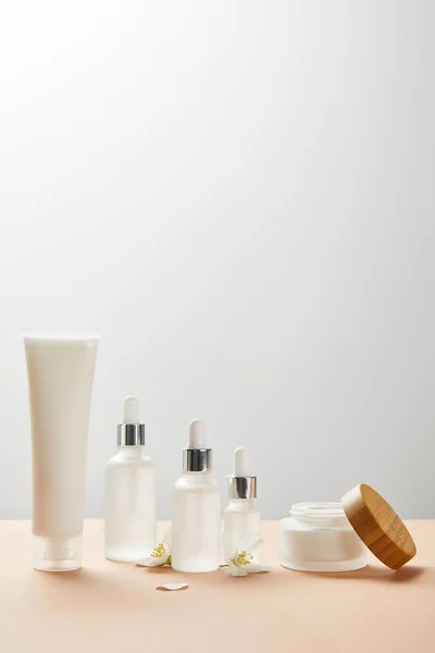 Tube à crème avec crème, bouteilles en verre cosmétique avec sérum, pot ouvert avec capuchon en bois et peu de fleurs de jasmin sur beige — Photo de stock