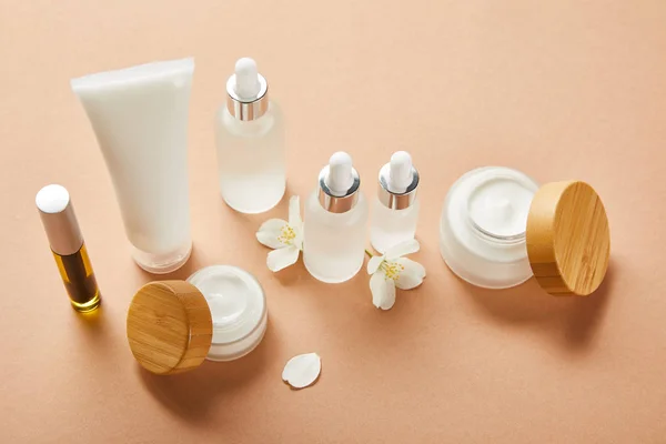 Bouteille de mascara avec huile naturelle, tube à crème, bouteilles en verre cosmétique, pots ouverts avec crème et fleurs de jasmin sur beige — Photo de stock