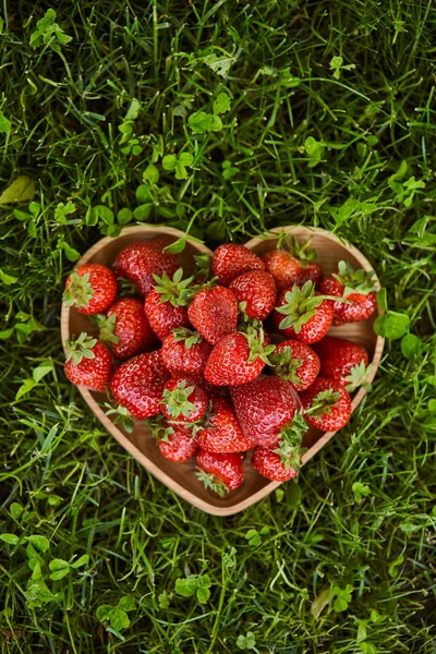 Vista dall'alto di fragole biologiche in piatto di legno a forma di cuore su erba verde — Foto stock