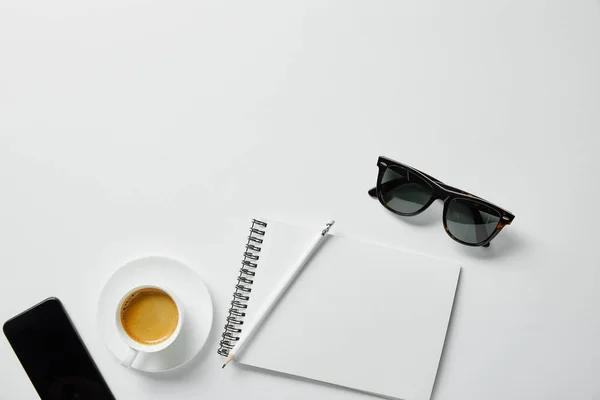 Vista dall'alto di caffè, notebook, matita e bicchieri con smartphone su superficie bianca — Stock Photo