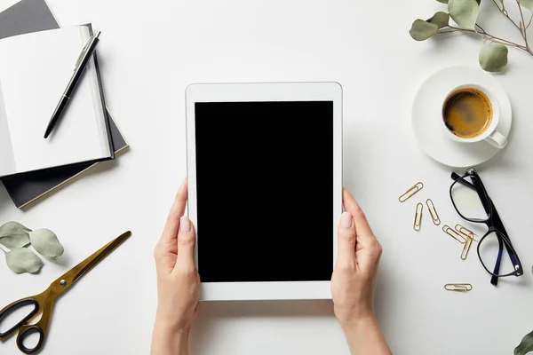 Vista recortada da mulher segurando tablet digital em mãos perto de plantas, artigos de papelaria, tesoura, café, óculos e cadernos com caneta na superfície branca — Fotografia de Stock