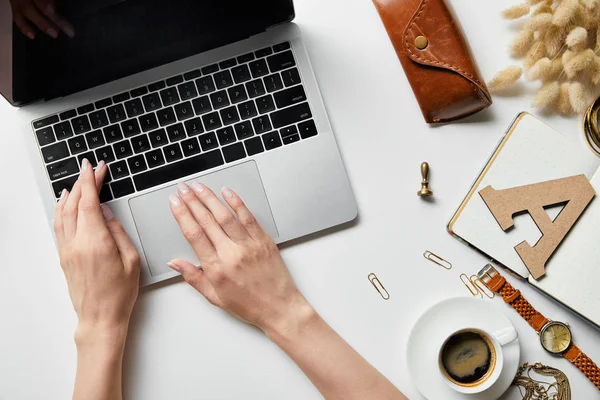 Vista ritagliata di donna che lavora con computer portatile su superficie bianca con piante, caffè, custodia, cancelleria e blocco note — Foto stock