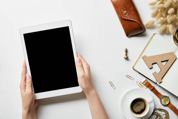 Vista recortada de la tableta digital en las manos de la mujer en la superficie blanca con papelería, joyas, bloc de notas, estuche y café - foto de stock