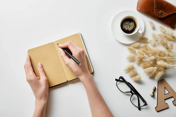 Cropped view of woman writing in notepad near coffee, glasses and case on white surface — Stock Photo