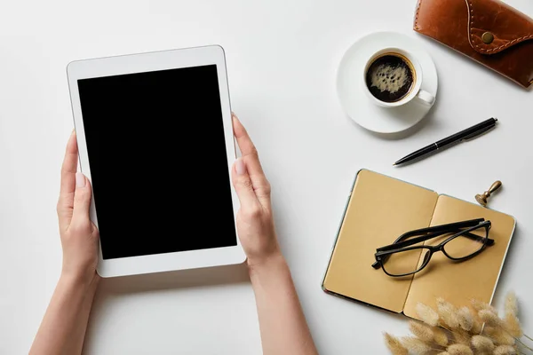 Vue recadrée de la femme tenant tablette numérique dans les mains près de bloc-notes, café, lunettes et étui sur la surface blanche — Photo de stock