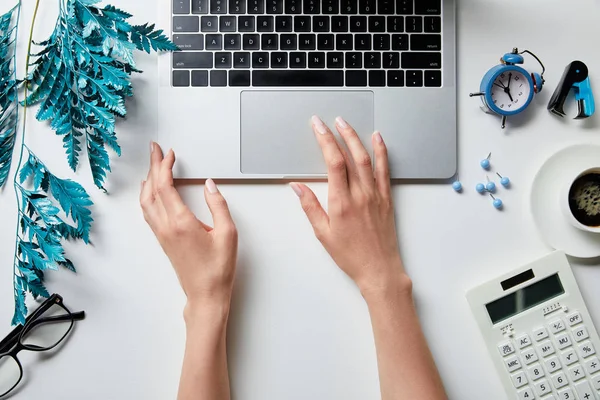 Vue recadrée de la femme travaillant avec un ordinateur portable près du café, papeterie, réveil, calculatrice, lunettes et branche bleue sur la surface blanche — Photo de stock