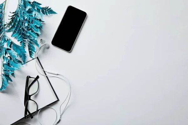 Vista superior del teléfono inteligente cerca del bloc de notas, gafas y rama azul en la superficie blanca - foto de stock