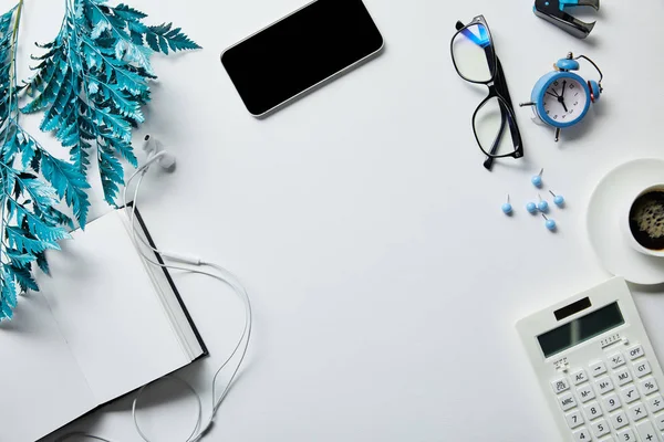 Vista superior del teléfono inteligente cerca del bloc de notas, café, papelería, auriculares, reloj despertador, calculadora, gafas y rama azul en la superficie blanca - foto de stock