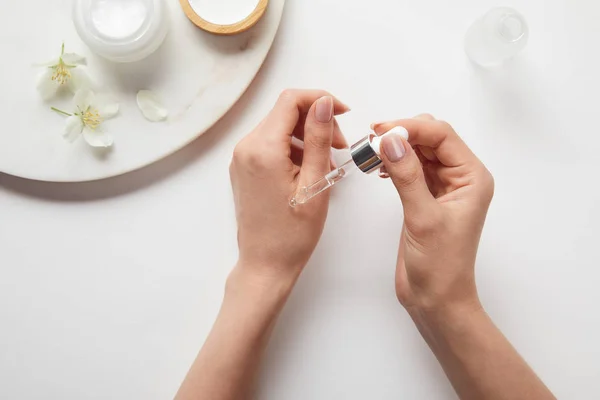 Vista recortada de la mujer aplicando líquido de botella de vidrio, cerca de la placa con crema y flores en la superficie blanca - foto de stock