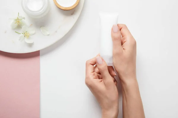 Vue recadrée de la femme tenant le tube de crème près de la plaque avec des cosmétiques et des fleurs sur la surface rose blanc — Photo de stock