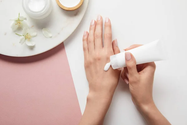 Visão recortada da mulher aplicando creme, segurando as mãos perto da placa com cosméticos e flores na superfície rosa branca — Fotografia de Stock