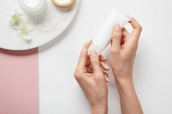 Vista recortada de la mujer sosteniendo tubo de crema cerca de la placa con cosméticos y flores en la superficie de color rosa blanco - foto de stock