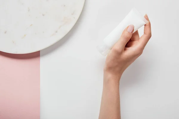 Vista recortada de la mujer sosteniendo tubo de crema cerca de la placa en la superficie de color rosa blanco - foto de stock