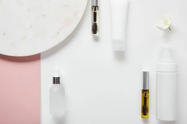 Vue du dessus du mascara et des bouteilles en verre, tube à crème, distributeur cosmétique près de la plaque, jasmin sur surface rose blanc — Photo de stock