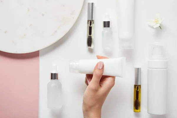 Cropped view of woman holding cream tube in hands over mascara and glass bottles, cosmetic dispenser, plate, jasmine on white pink surface — Stock Photo