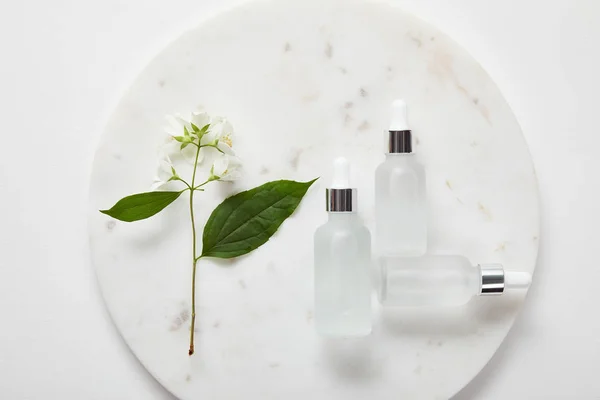 Vista dall'alto della lastra con gelsomino e bottiglie di vetro cosmetico su superficie bianca — Foto stock