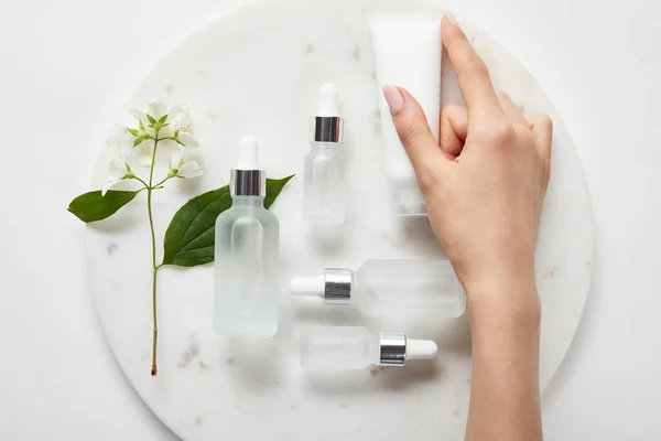 Abgeschnittene Ansicht der Frauenhand mit Cremetubus über Teller mit Jasmin- und Kosmetikglasflaschen auf weißer Oberfläche — Stockfoto