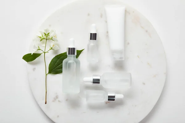 Vue du dessus du tube à crème et des bouteilles en verre cosmétique en plaque avec du jasmin sur surface blanche — Photo de stock