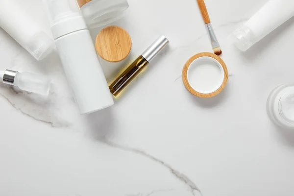 Cropped view of cream tubes, cosmetics jars, dispenser and glass bottle with eye brush on white surface — Stock Photo