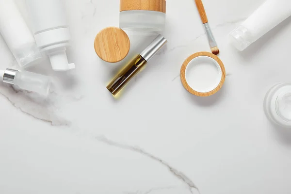 Cropped view of cream tubes, cosmetics jars, dispenser and glass bottle with eye brush on white surface — Stock Photo