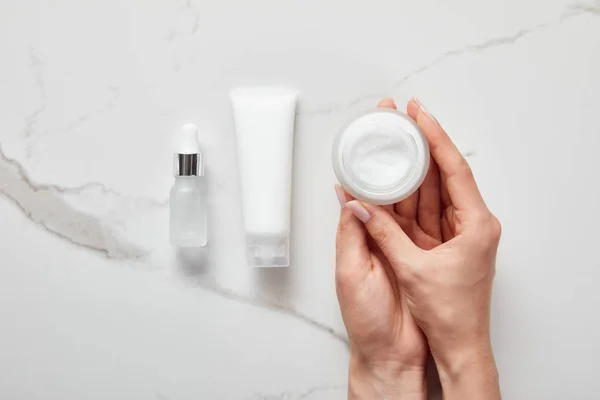 Top view of woman holding jar with cream near cosmetic glass bottle and moisturizer tube on white surface — Stock Photo