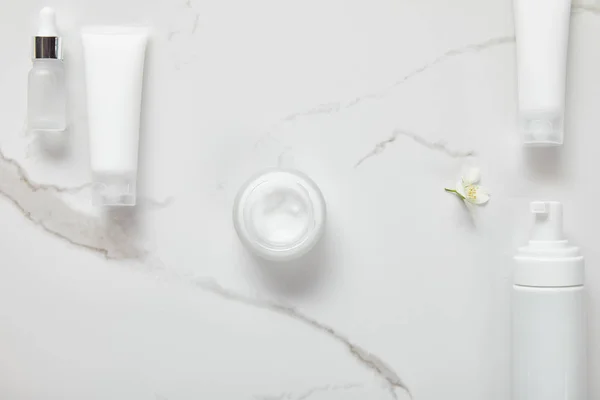 Top view of cosmetic glass bottle, jar with cream, moisturizer tubes, dispenser and jasmine on white surface — Stock Photo