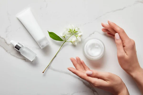 Vista dall'alto delle mani della donna vicino alla bottiglia di vetro cosmetico, vaso con crema, tubo idratante e gelsomino sulla superficie bianca — Foto stock