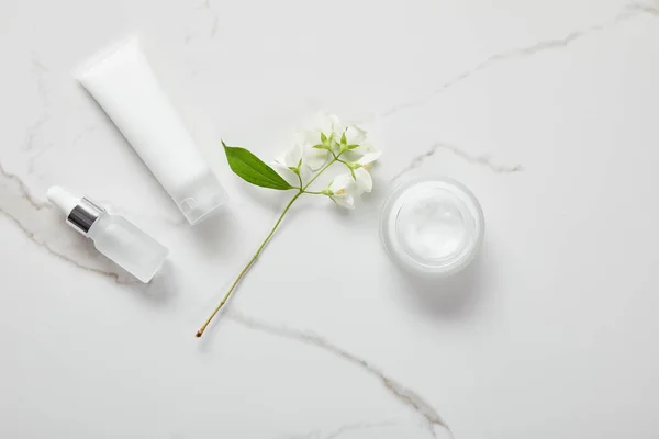 Top view of cosmetic glass bottle, jar with cream, moisturizer tube and jasmine on white surface — Stock Photo