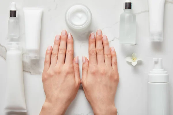 Vista ritagliata di mani di donna vicino a bottiglie di vetro cosmetico, vaso con crema, tubi idratanti, dispenser e gelsomino sulla superficie bianca — Foto stock