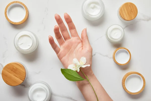 Vista recortada de flores de jazmín en la mano de la mujer cerca de frascos con crema cosmética en la superficie blanca — Stock Photo