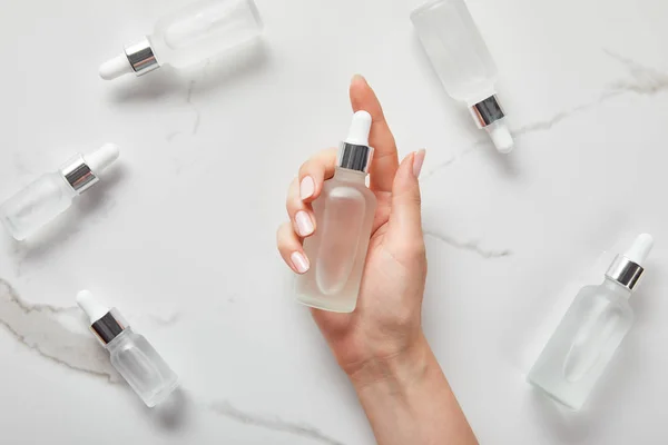 Vista cortada da mão da mulher com soro hidratante perto de garrafas de vidro cosmético na superfície branca — Fotografia de Stock