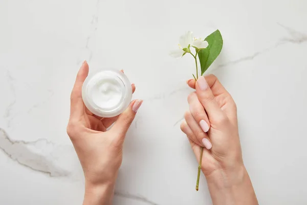 Vista cortada da mulher segurando flores de jasmim e frasco com creme sobre a superfície branca — Fotografia de Stock