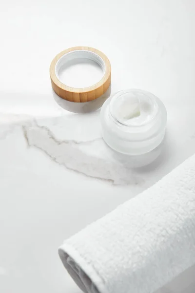 Pot ouvert avec crème cosmétique et capuchon en bois près de la serviette sur la surface blanche — Photo de stock