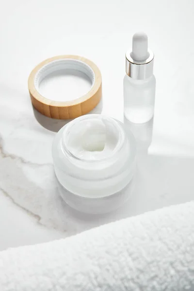 Frasco abierto con crema cosmética y botella de vidrio cerca de la toalla en la superficie blanca - foto de stock