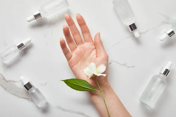 Vue recadrée de la main de la femme avec du jasmin près des bouteilles en verre cosmétique sur la surface blanche — Photo de stock