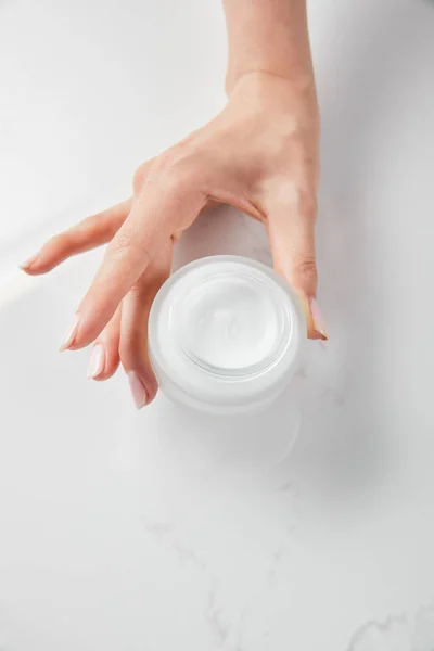 Vista recortada de la mujer sosteniendo tarro con crema sobre la superficie blanca - foto de stock