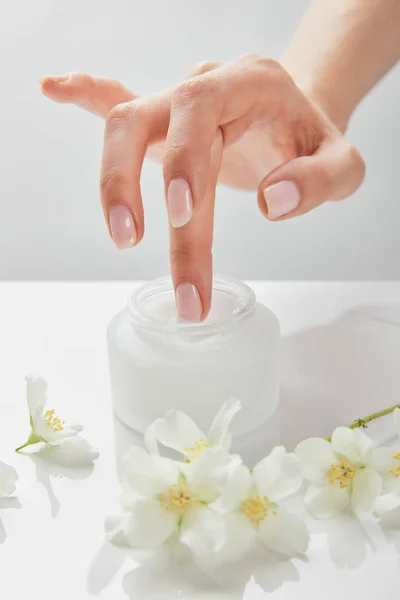 Vista recortada de la mano de la mujer tocando crema en tarro cerca de flores de jazmín en la superficie blanca - foto de stock