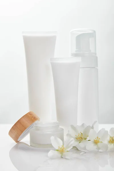 Flores de jazmín en la superficie blanca cerca de la crema en tubos, frasco y dispensador de cosméticos - foto de stock